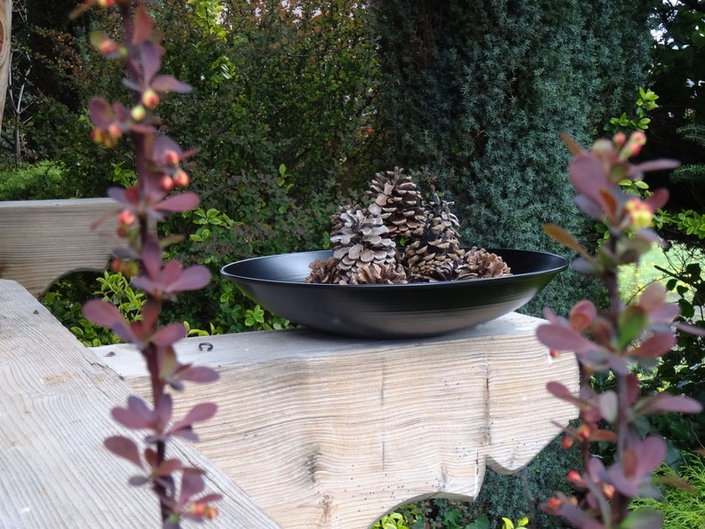 Op de afbeelding: Een zwarte kom gevuld met dennenappels staat op een houten oppervlak. De kom is omringd door groen gebladerte en een tak met rode bladeren.