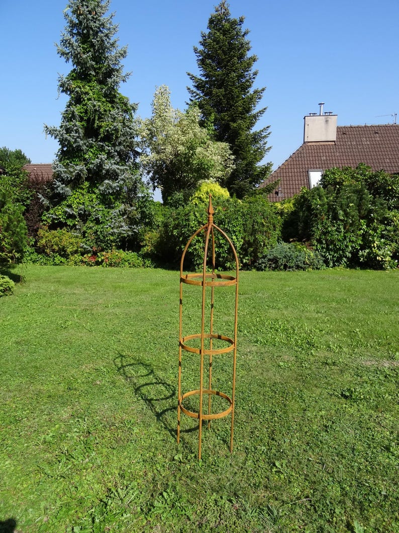 Op de afbeelding: Een hoge, cilindrische tuinobelisk van roestig metaal. De structuur is voorzien van een puntige top, drie cirkelvormige horizontale ringen verbonden door verticale stangen en vier poten die naar beneden uitsteken voor in de grond.