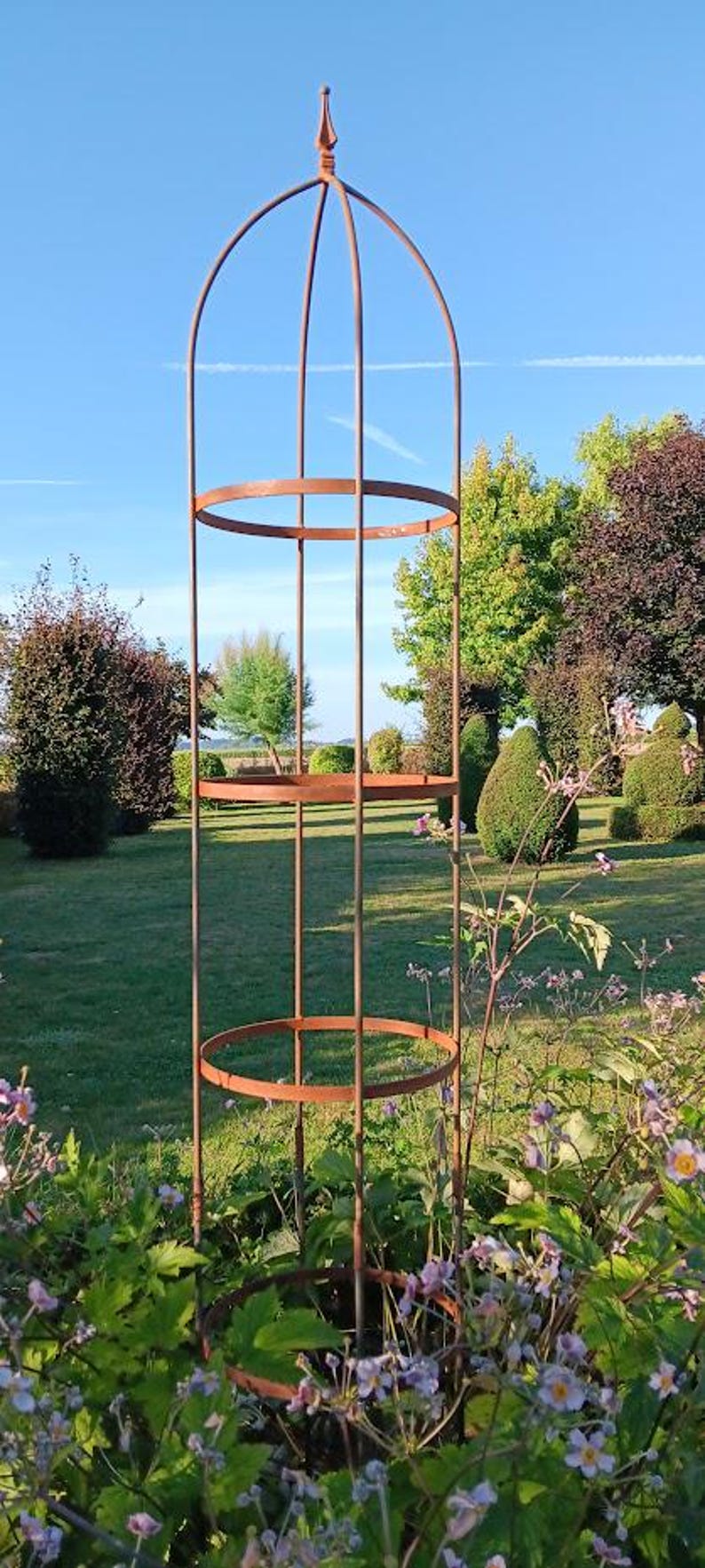 Op de afbeelding: Een hoge, roestkleurige metalen tuinobelisk met een verticaal frame, drie cirkelvormige steunringen en een puntige knop aan de bovenkant. De structuur is ontworpen voor klimplanten en staat rechtop in een tuin.