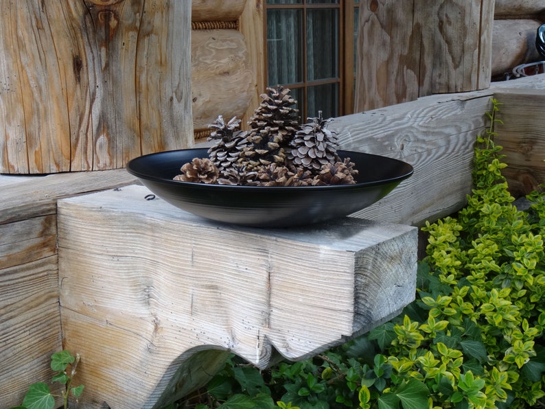 Op de afbeelding: Een zwarte keramische kom gevuld met dennenappels staat op een houten balk. De kom is rond en heeft een licht verhoogde rand. De dennenappels zijn bruin en hebben een natuurlijke, rustieke uitstraling.