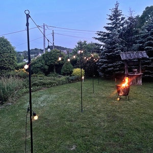 Puede incluir: Una cadena de luces colgadas entre postes de metal negro en un jard&iacute;n. Las luces est&aacute;n tendidas sobre una zona de c&eacute;sped con una hoguera en el centro.