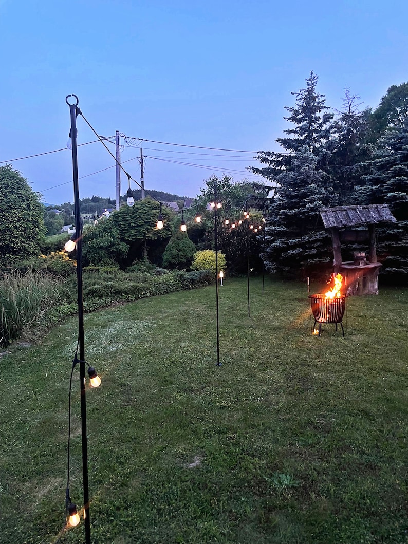 Puede incluir: Una cadena de luces colgadas entre postes de metal negro en un jard&iacute;n. Las luces est&aacute;n tendidas sobre una zona de c&eacute;sped con una hoguera en el centro.