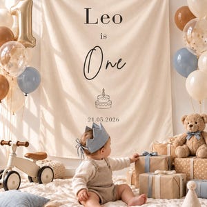 May include: A cream-coloured fabric wall hanging featuring the text Leo is One, a line-art illustration of a birthday cake, and the date 21.05.2026. The banner is displayed in a room decorated with balloons, a teddy bear, and gifts.