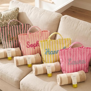 May include: Several striped tote bags in pink, green, and yellow, each personalised with a name in a contrasting colour. Beach towels and sunscreen tubes are arranged in front of the bags on a beige sofa.