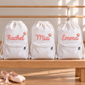 Könnte beinhalten: Drei weiße Rucksäcke mit Kordelzug und strukturiertem Rautenmuster, jeweils mit einer Reißverschlusstasche auf der Vorderseite und einem in rosa Schreibschrift gestickten Namen: Rachel, Mia und Emma, ergänzt durch eine kleine rosa Blume.