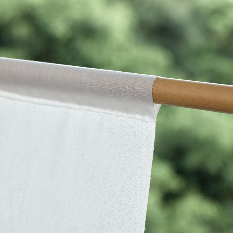 May include: Close-up of a white curtain panel attached to a light brown wooden rod. The curtain is made of a sheer, lightweight fabric. The background is a blurred view of green foliage.
