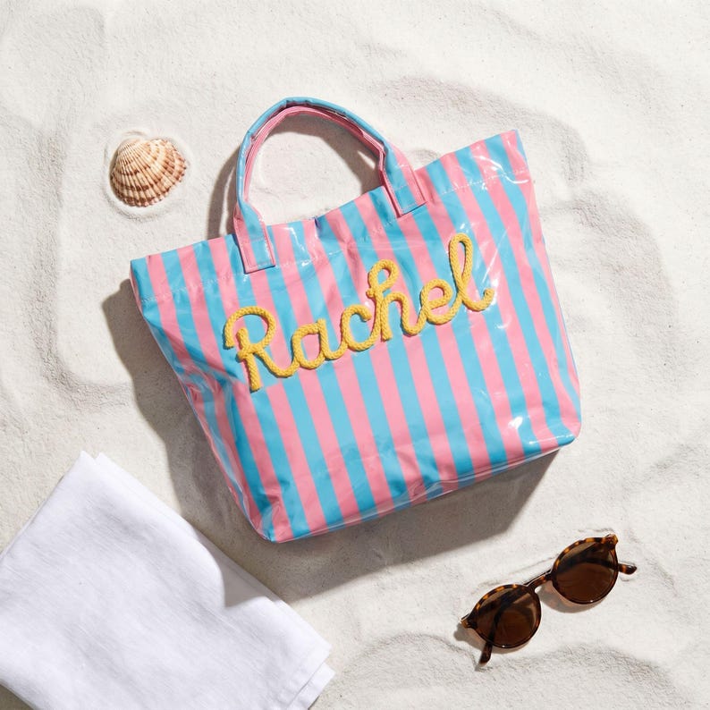 May include: A pink and blue striped tote bag with the name "Rachel" embroidered in yellow. The bag has matching pink and blue handles. A seashell, sunglasses, and a white towel are also on the sandy surface.