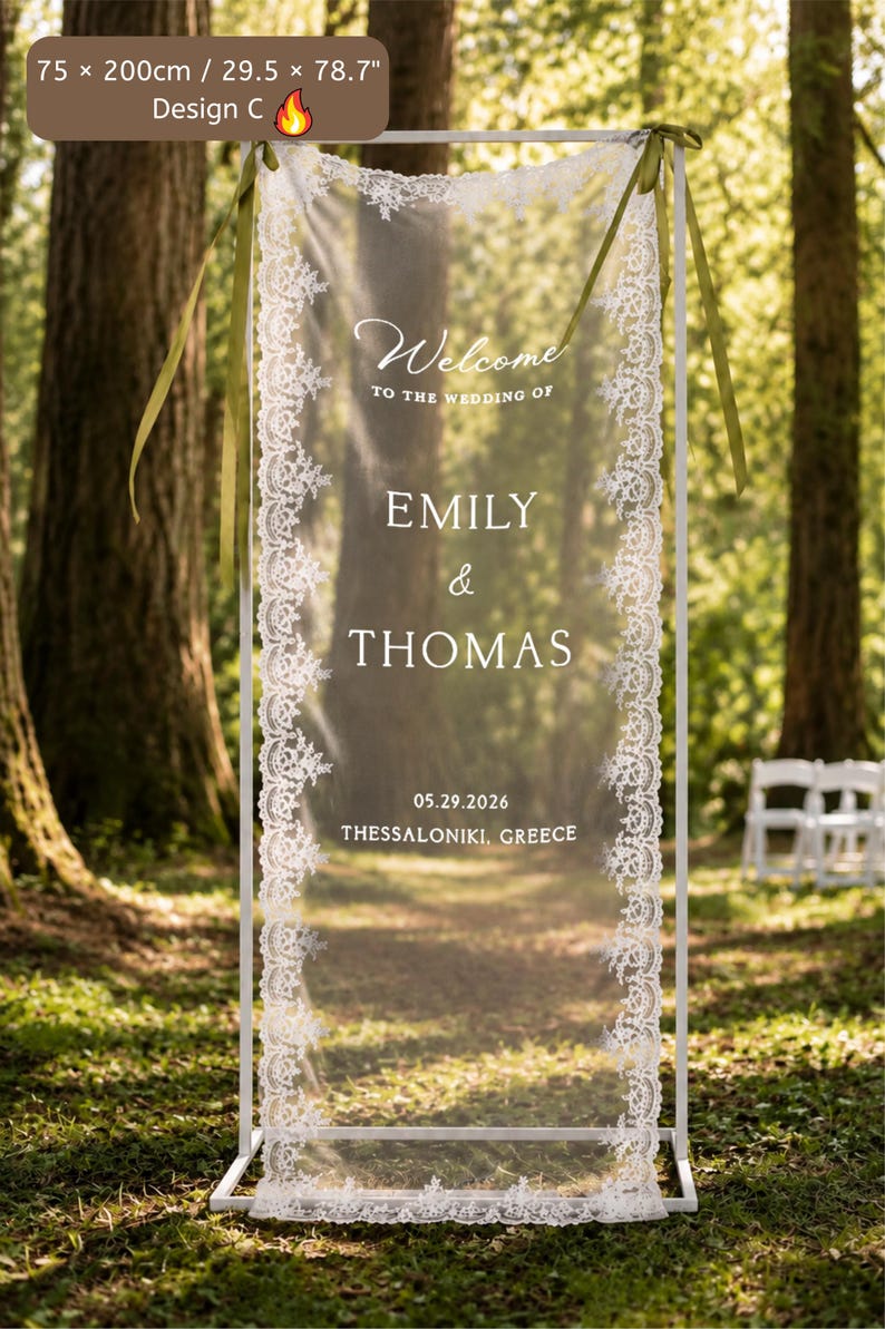 Puede incluir: Un marco met&aacute;lico blanco rectangular que sostiene un panel de tela transparente con borde de encaje blanco. El panel presenta el texto Welcome to the wedding of Emily & Thomas 05.29.2026 Thessaloniki, Greece. Cintas verde oliva en las esquinas.