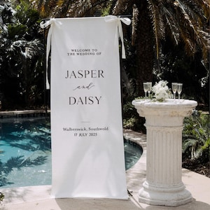May include: A white wedding banner with the text "WELCOME TO THE WEDDING OF JASPER and DAISY" and the date "17 JULY 2025". The banner is 29.5 x 78.7 inches. A decorative pillar holds champagne glasses and flowers.