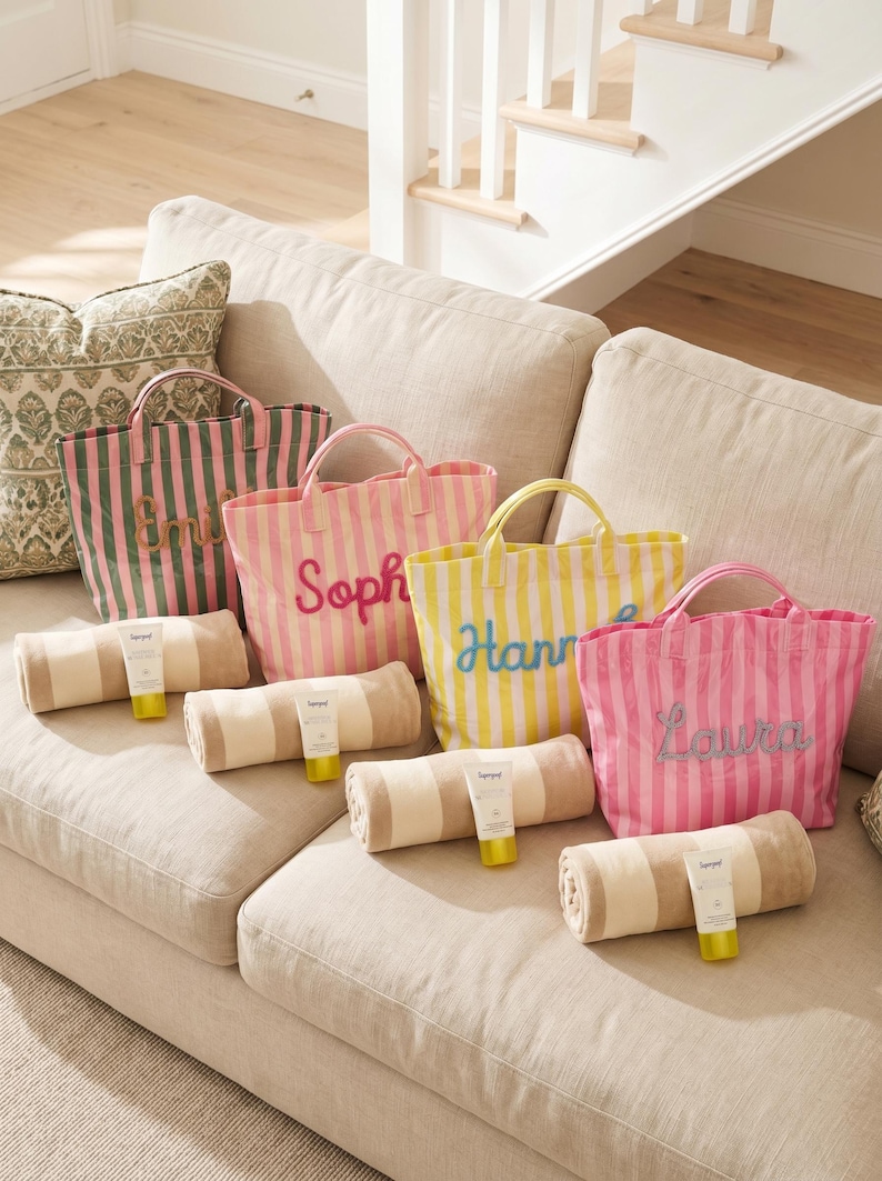 May include: Several striped tote bags in pink, green, and yellow, each personalised with a name in a contrasting colour. Beach towels and sunscreen tubes are arranged in front of the bags on a beige sofa.