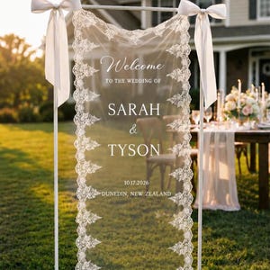 Puede incluir: Un letrero de bienvenida de boda rectangular de 65 x 165 cm, con un panel de tela transparente con borde de encaje blanco, suspendido de un marco de metal blanco con lazos de cinta blanca. Texto: Welcome TO THE WEDDING OF SARAH & TYSON 10.17.2026 DUNEDIN, NEW ZEALAND.