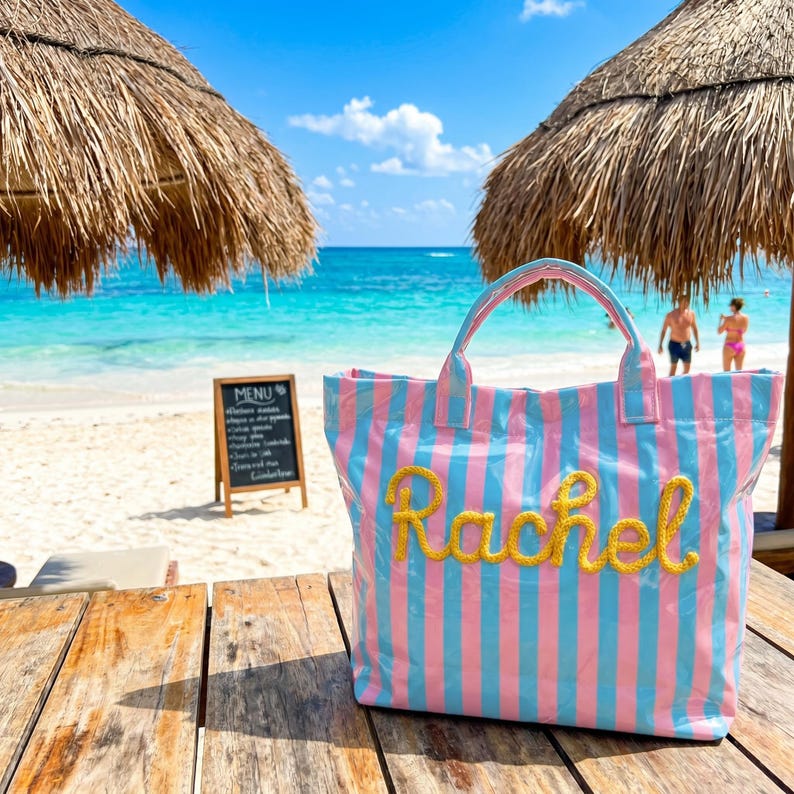 May include: A pink and blue striped tote bag with the name "Rachel" in gold glitter lettering. The bag is on a wooden table with a beach and ocean in the background. A menu board and thatched umbrellas are also visible.