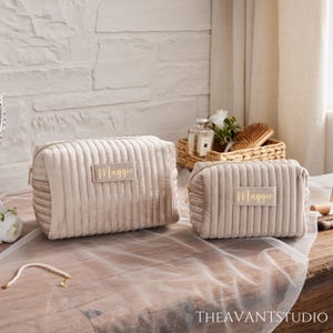 Peut inclure: Deux trousses de maquillage en velours côtelé beige avec des fermetures éclair dorées et le nom "Maggie" sur une étiquette rectangulaire. Les sacs sont de tailles différentes et reposent sur une surface en bois. L'arrière-plan comprend un mur de briques blanches et un rideau blanc transparent.