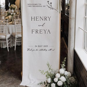 May include: A wedding welcome sign with the text "WELCOME TO THE WEDDING OF HENRY and FREYA" and the date "26 JULY 2025". The sign is made of a cream-colored fabric and is 29.5 inches wide and 78.7 inches tall. Floral arrangements are at the base.