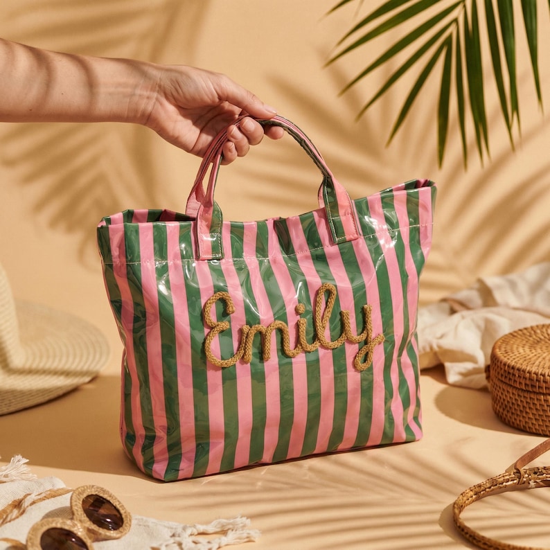 May include: A pink and green striped tote bag with the name "Emily" embroidered in a rope-like material. The bag has a glossy finish and sturdy handles. The bag is a stylish accessory for carrying essentials.