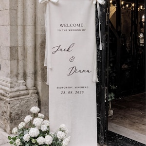 May include: A cream-colored wedding banner with the text "WELCOME TO THE WEDDING OF Jack & Diana" and the date 25.08.2025. The banner is 27.6 x 78.7 inches and is tied with white ribbons. White flowers are at the base.