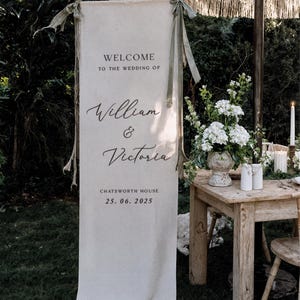 May include: A cream-colored wedding welcome sign with the text "WELCOME TO THE WEDDING OF William & Victoria" and the date 25.06.2025. The sign is 27.6 x 78.7 inches. The sign is hanging outdoors with a decorated table and flowers.