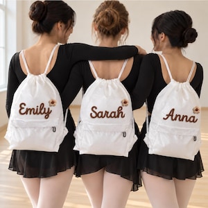 以下が含まれることがあります： Emily、Sarah、Annaという名前が茶色で刺繍された3つの白い巾着型バックパック。各バッグには名前の近くに小さな茶色の花柄のアップリケ、黒い金具が付いたフロントジッパーポケット、白いコードストラップが付いています。