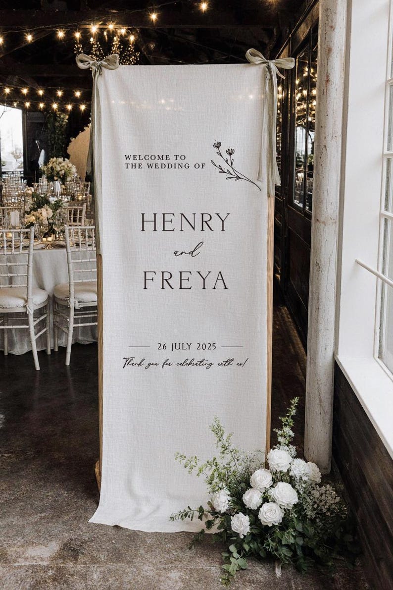 May include: A white linen wedding sign with the text "WELCOME TO THE WEDDING OF HENRY and FREYA 26 JULY 2025". The sign is decorated with floral accents and greenery. The sign is propped up with wooden supports.
