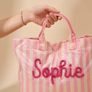 May include: A pink and white striped tote bag with the name "Sophie" embroidered in pink yarn. The bag has pink handles and is held by a person. The bag is made of a clear, shiny material.