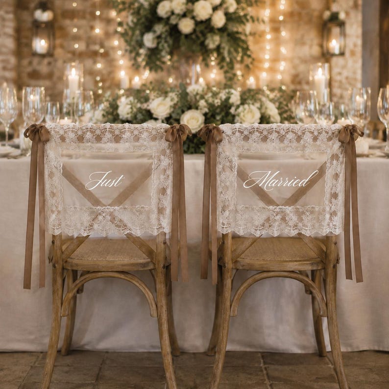 Puede incluir: Dos sillas de madera con fundas de encaje blanco y lazos marrones. La funda de la izquierda muestra el texto blanco Just, y la de la derecha el texto blanco Married. El encaje presenta un patr&oacute;n floral y bordes con acabado festoneado.