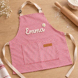 May include: A red and white vertically striped apron featuring the name Emma in white script lettering, a small white flower embellishment, and a rectangular front pocket with a brown leather-like tag labeled with the word apron in lowercase.