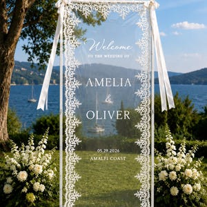 Puede incluir: Un letrero de bienvenida para bodas alto, rectangular y transparente de acr&iacute;lico que mide 75 x 200 cm. Presenta un borde de encaje blanco, cintas blancas en las esquinas superiores y texto blanco que dice Welcome TO THE WEDDING OF AMELIA & OLIVER 29.05.2026 AMALFI COAST.