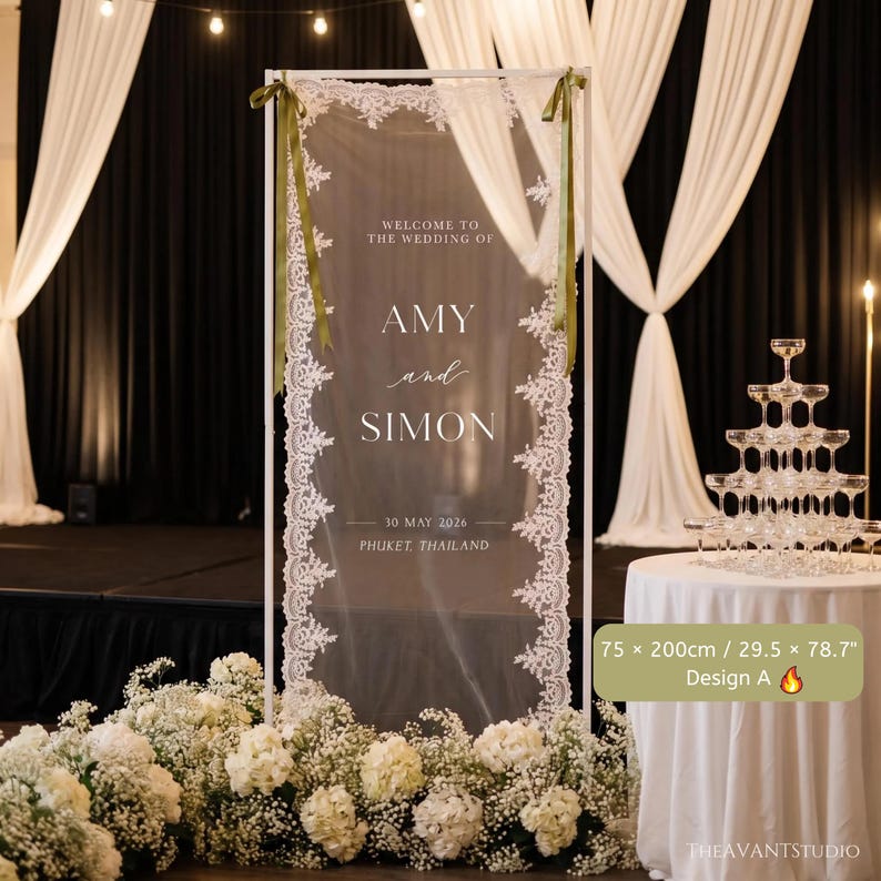 Puede incluir: Un letrero de bienvenida de boda rectangular con un panel de tela transparente y bordes de encaje blanco, sostenido por un marco blanco con cintas verde oliva. El texto dice: WELCOME TO THE WEDDING OF AMY and SIMON 30 MAY 2026 PHUKET, THAILAND. Mide 75 x 200 cm.