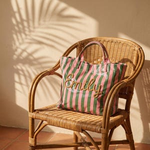 May include: A pink and green striped tote bag with the name "Emily" embroidered on the front. The bag is on a wicker chair. The background shows shadows on a wall.