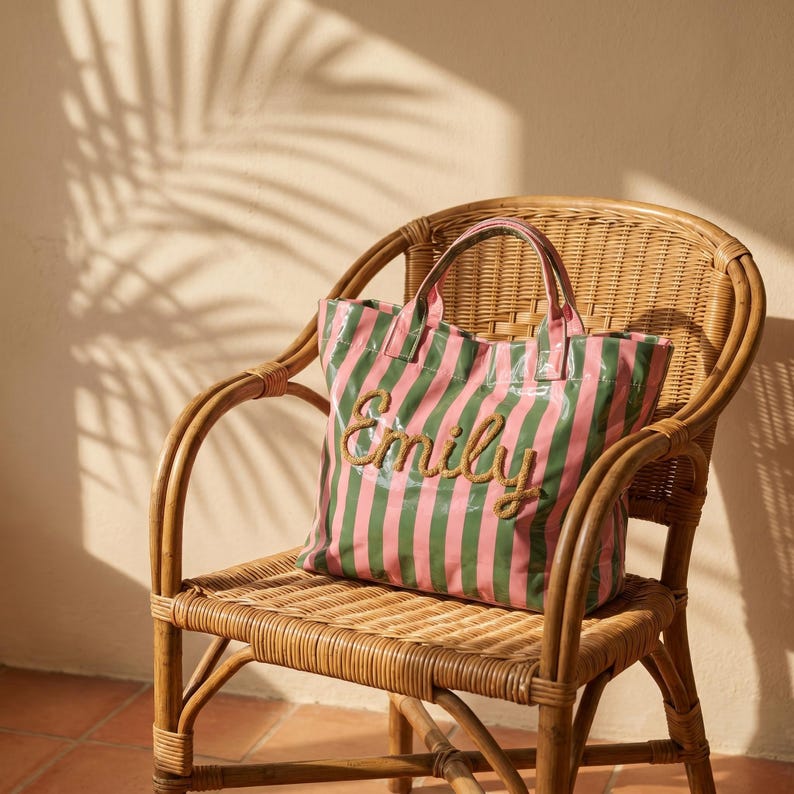 May include: A pink and green striped tote bag with the name "Emily" embroidered on the front. The bag is on a wicker chair. The background shows shadows on a wall.