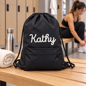 Könnte beinhalten: Ein schwarzer Kordelzugrucksack mit texturiertem Rautenmuster und dem Namen Kathy, der in weißer Schreibschrift auf der Vorderseite eingestickt ist. Die Tasche verfügt über eine Reißverschlusstasche vorne und dicke schwarze Kordelträger.