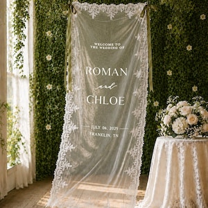 Puede incluir: Un letrero de boda de tela blanca transparente con bordes de encaje intrincados, suspendido por cintas de color verde oliva. El texto dice: WELCOME TO THE WEDDING OF ROMAN and CHLOE JULY 06, 2025 FRANKLIN, TN. Presenta un delicado patr&oacute;n de encaje floral.