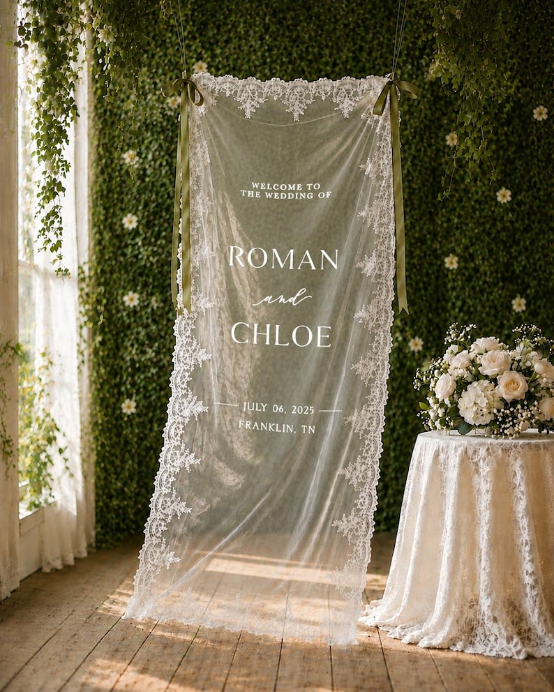 Puede incluir: Un letrero de boda de tela blanca transparente con bordes de encaje intrincados, suspendido por cintas de color verde oliva. El texto dice: WELCOME TO THE WEDDING OF ROMAN and CHLOE JULY 06, 2025 FRANKLIN, TN. Presenta un delicado patr&oacute;n de encaje floral.