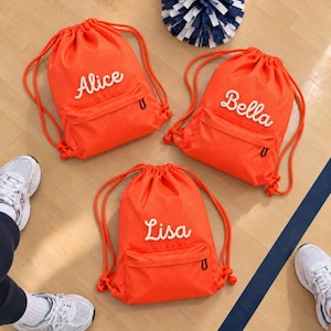 Puede incluir: Tres mochilas de cordón de color naranja brillante hechas de material de malla texturizada. Cada bolsa cuenta con un bolsillo frontal con cremallera y un nombre bordado en blanco: Alice, Bella o Lisa. Las bolsas tienen cordones naranjas.