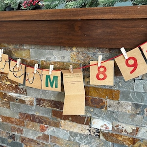 May include: A handmade advent calendar with brown paper squares numbered 8, 9, and 10 in red, strung on a red cord. The squares are clipped to the cord with small wooden clothespins. The background is a stone wall.
