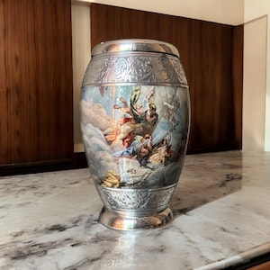 May include: A silver vase with an ornate design and a painted scene of figures in the clouds. The vase has a round body and a wide base.