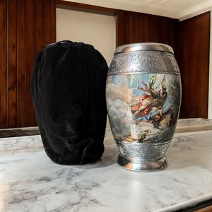 May include: A silver urn with an ornate design and a painted scene of angels in the clouds. The urn is sitting on a marble surface next to a black velvet bag.