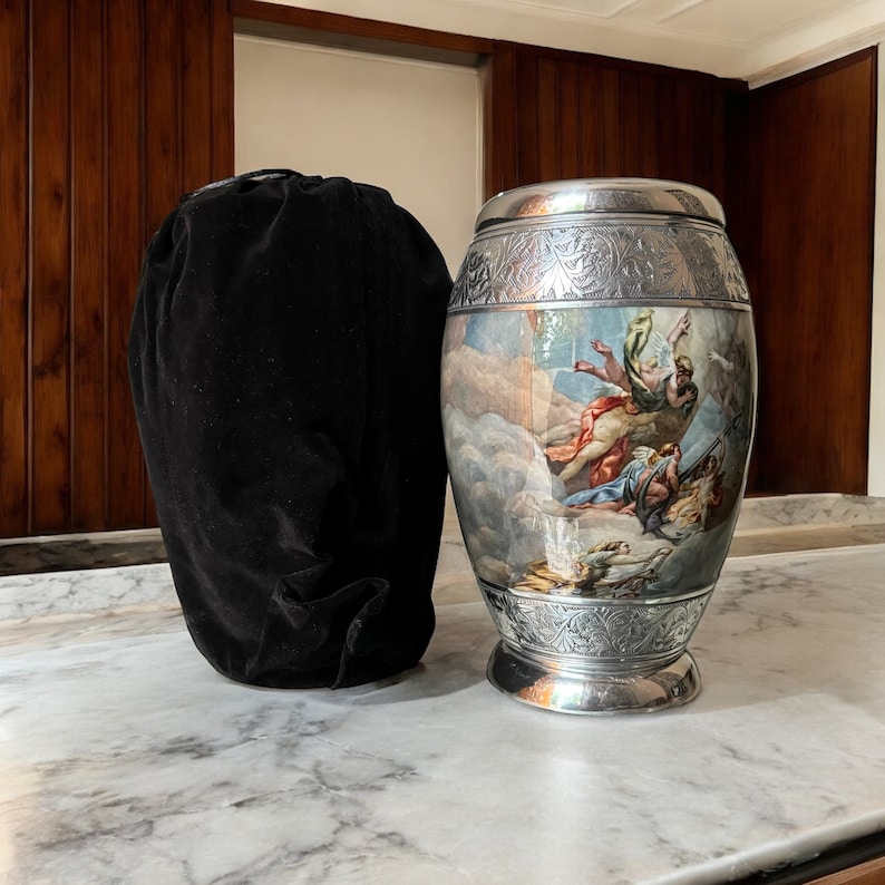 May include: A silver urn with an ornate design and a painted scene of angels in the clouds. The urn is sitting on a marble surface next to a black velvet bag.