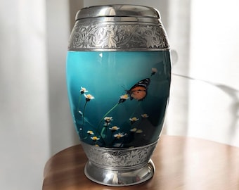 May include: A silver urn with an engraved floral design and a blue background featuring a butterfly and white flowers.