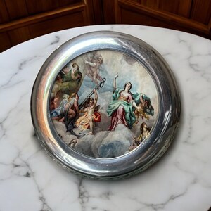 May include: A silver circular trinket box with a raised rim and a painted scene of a woman and angels on clouds. The scene is painted in shades of blue, green, pink, and yellow.