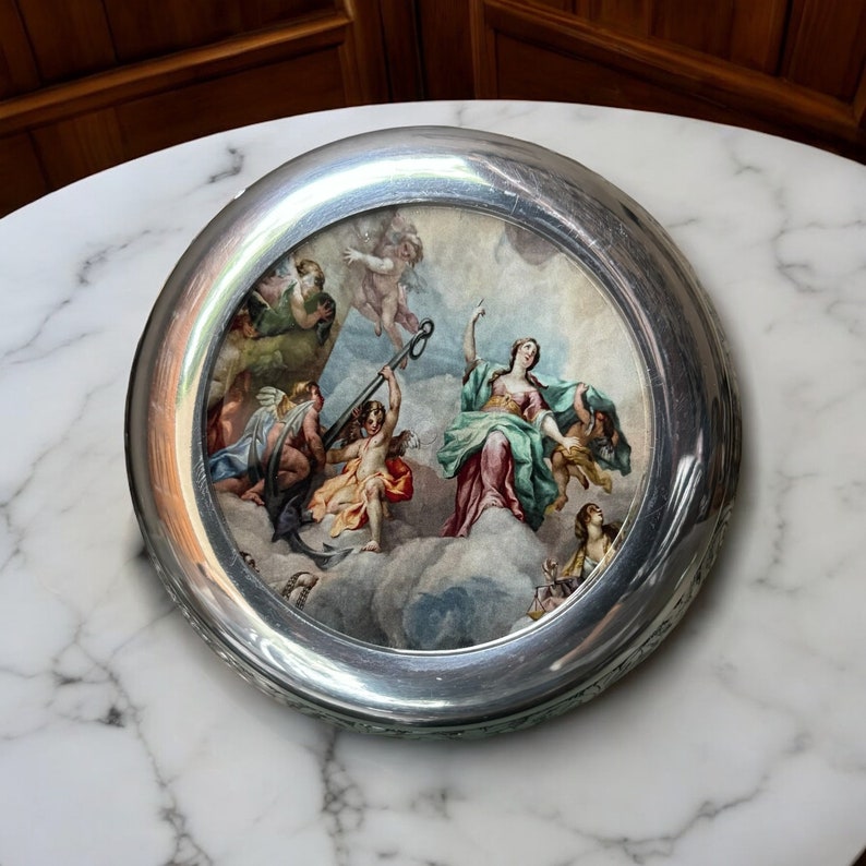 May include: A silver circular trinket box with a raised rim and a painted scene of a woman and angels on clouds. The scene is painted in shades of blue, green, pink, and yellow.