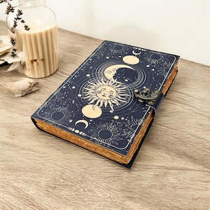 May include: A dark blue journal with a celestial design featuring a sun, moon, and stars. The journal has a metal clasp and aged-looking pages. The cover design is in cream and gold tones.