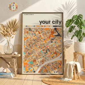 Puede incluir: Impresión enmarcada de un mapa de la ciudad con el texto "your city". El mapa presenta una representación abstracta del diseño de la ciudad, con un río. La paleta de colores incluye tonos de naranja, marrón y beige. La impresión se muestra en una habitación con plantas y muebles de madera.