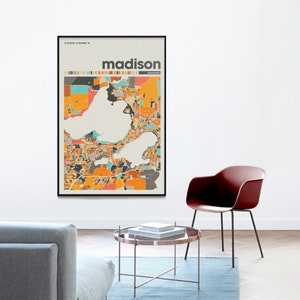 May include: A framed print of a colorful map of Madison, Wisconsin. The map is in a black frame and the city name is printed in bold, black letters at the top of the map. The map is hanging on a white wall above a brown coffee table and a red chair.