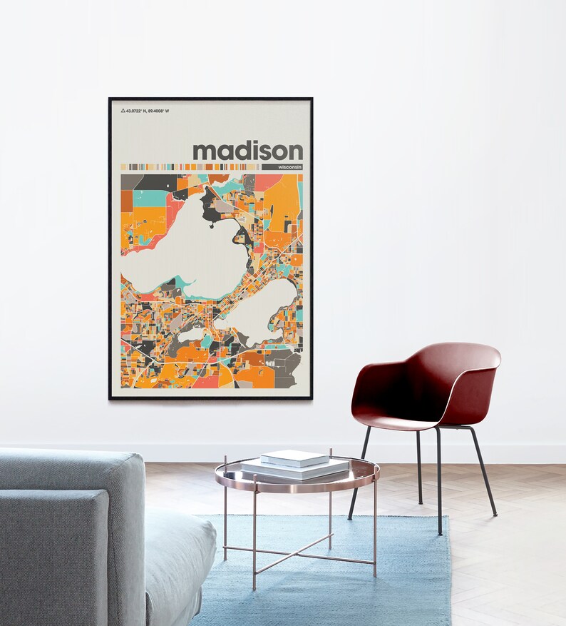 May include: A framed print of a colorful map of Madison, Wisconsin. The map is in a black frame and the city name is printed in bold, black letters at the top of the map. The map is hanging on a white wall above a brown coffee table and a red chair.