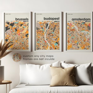 May include: Three framed city maps with a colorful, abstract style. The maps are of Brussels, Budapest, and Amsterdam. The frames are white and the maps are printed on white paper. The maps are hanging on a white wall above a white couch with beige pillows.