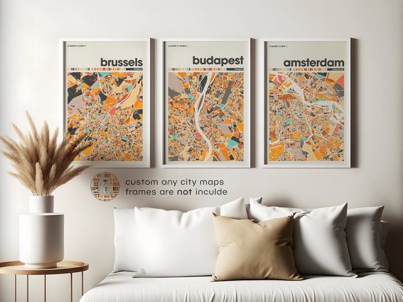 May include: Three framed city maps with a colorful, abstract style. The maps are of Brussels, Budapest, and Amsterdam. The frames are white and the maps are printed on white paper. The maps are hanging on a white wall above a white couch with beige pillows.