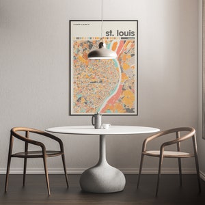 May include: A white table with a gray base and two wooden chairs sits in a room with a light gray wall. A black framed poster with a colorful map of St. Louis hangs on the wall above the table. A gray pendant light hangs above the table.