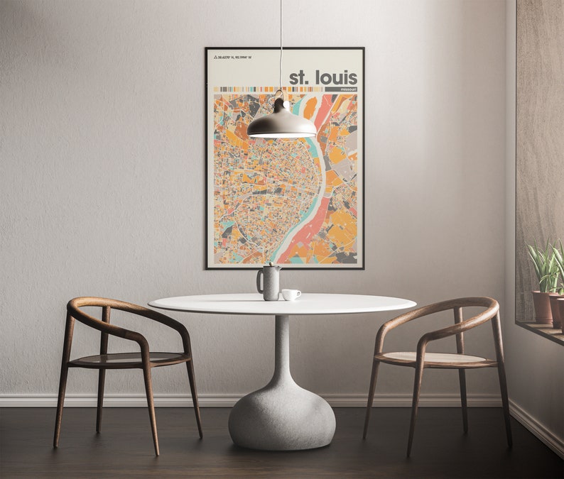 May include: A white table with a gray base and two wooden chairs sits in a room with a light gray wall. A black framed poster with a colorful map of St. Louis hangs on the wall above the table. A gray pendant light hangs above the table.