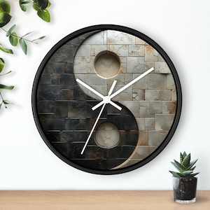 Pode incluir: Relógio de parede redondo com moldura preta. O mostrador apresenta um design Yin e Yang, com um padrão texturizado de azulejos quadrados em preto e cinza de um lado e bege e cinza do outro. Os ponteiros brancos indicam as horas.